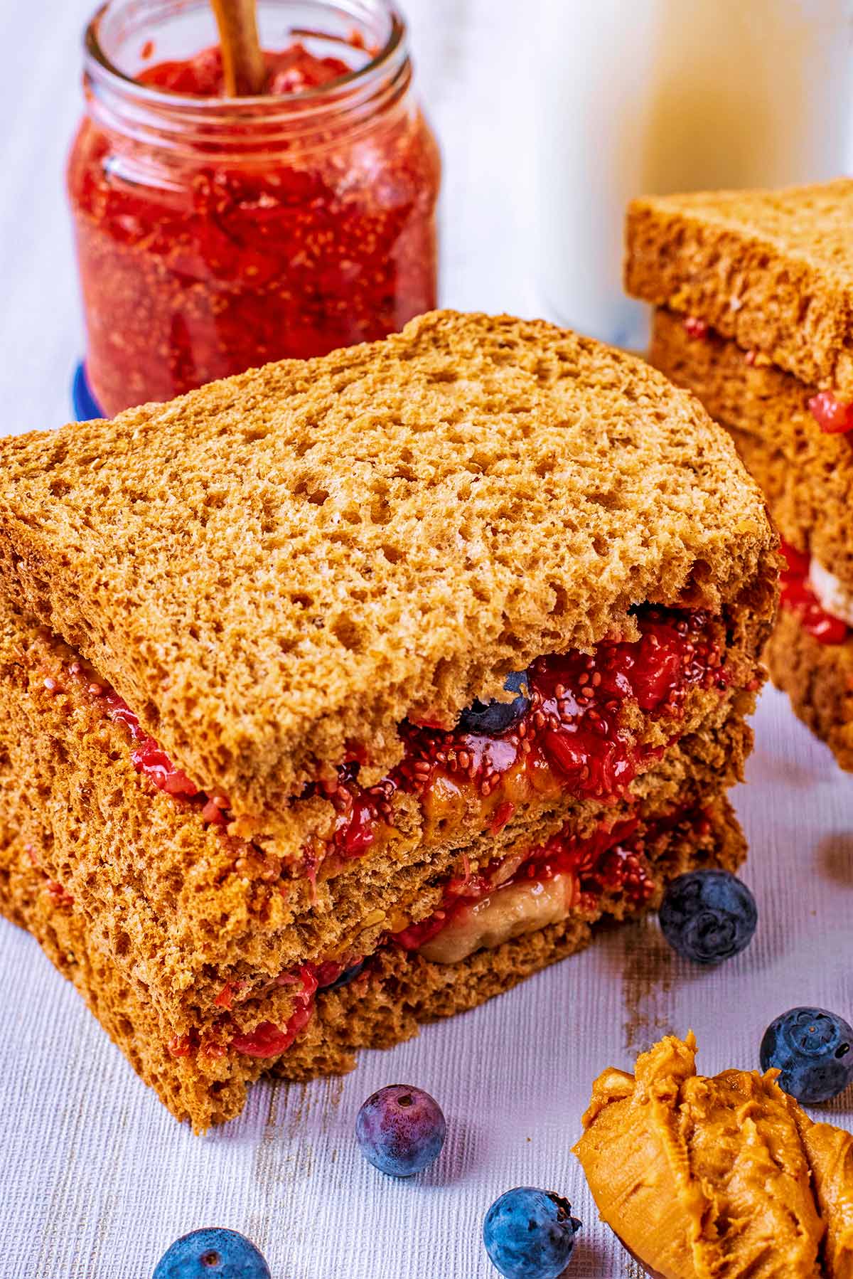 Two sandwiches stacked in front of a pot of jam and bottle of milk.