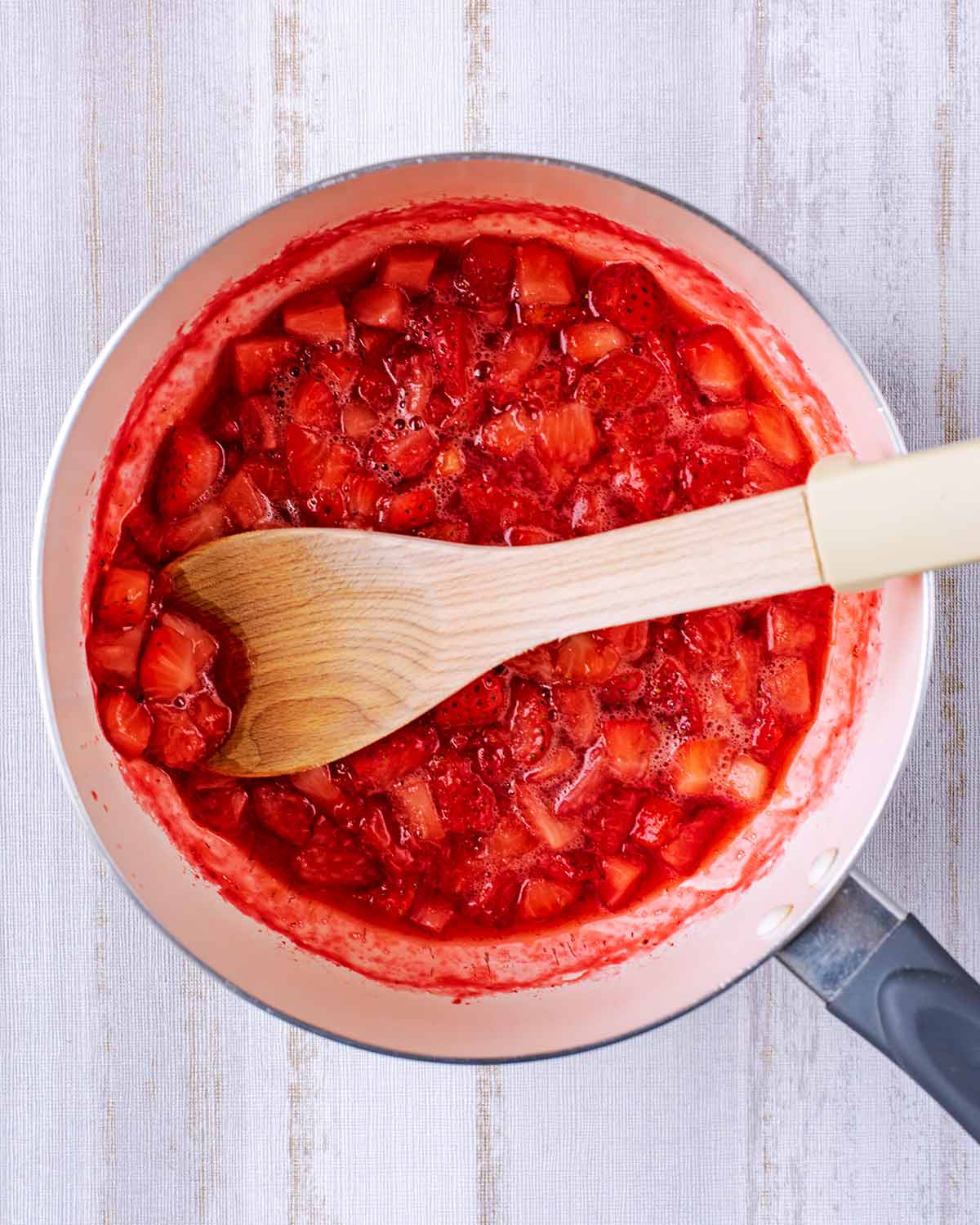 A wooden spoon mashing cooked strawberries in a pan.