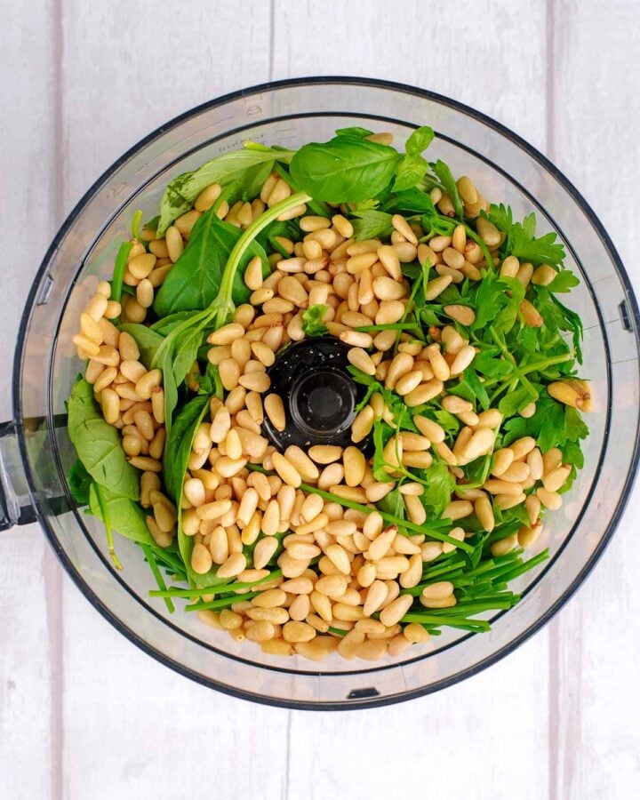 A food processor with herbs and pine nuts in it.
