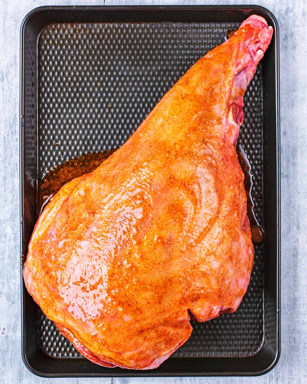 A leg of lamb covered in marinade on a black baking tray.