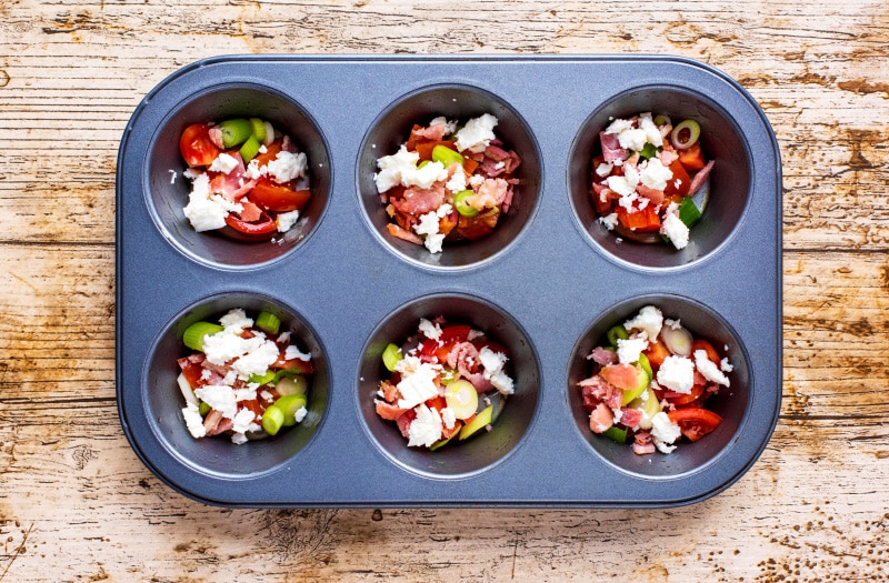 A six hole muffin tin with each hole containing dome red pepper, bacon, onion and cheese.