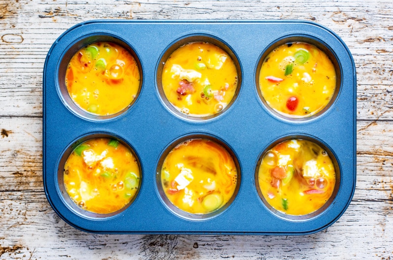 A six hole muffin tin with each hole containing beaten egg, chopped vegetables and cheese.