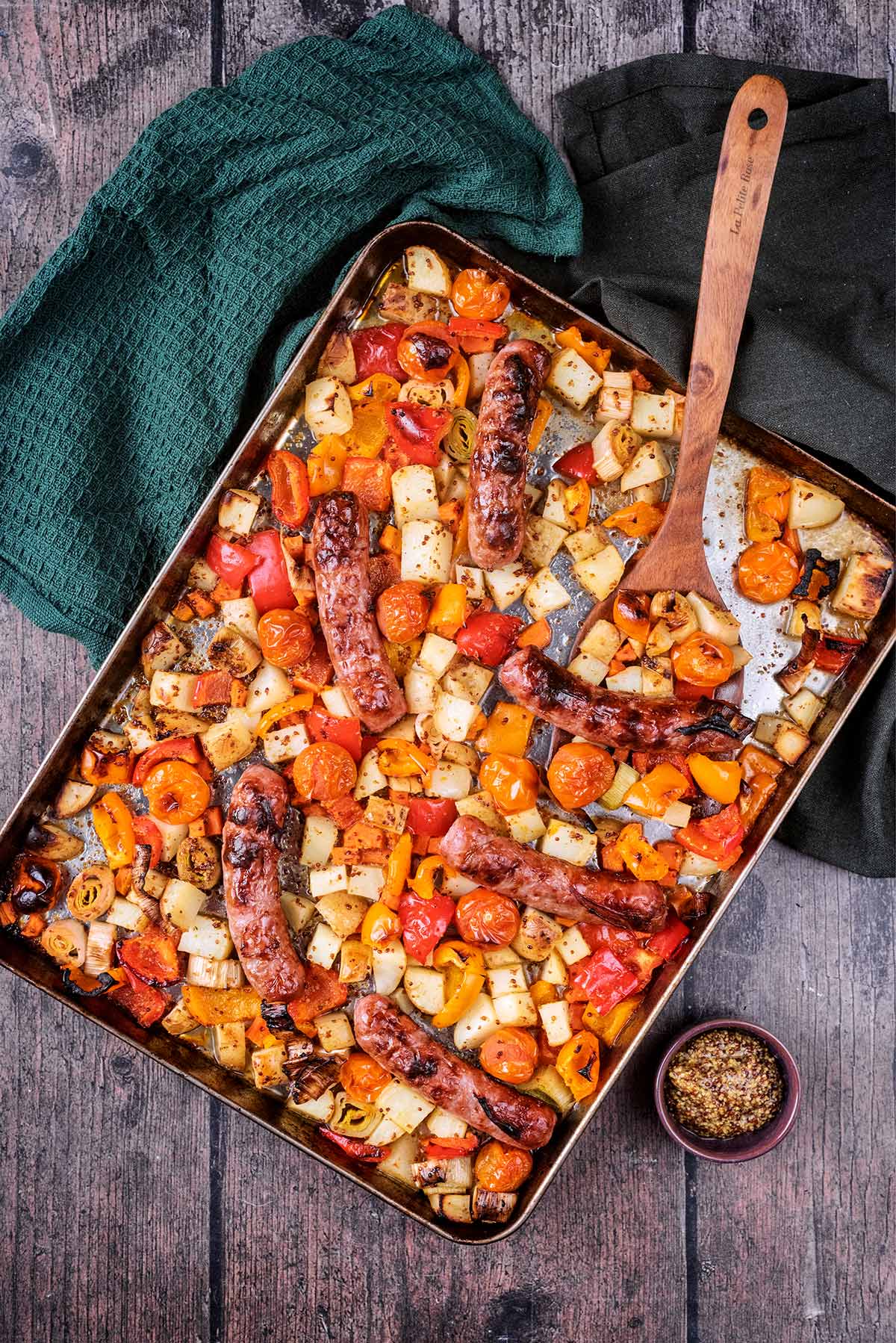 A traybake of sausages and vegetables with a large wooden spoon in it.