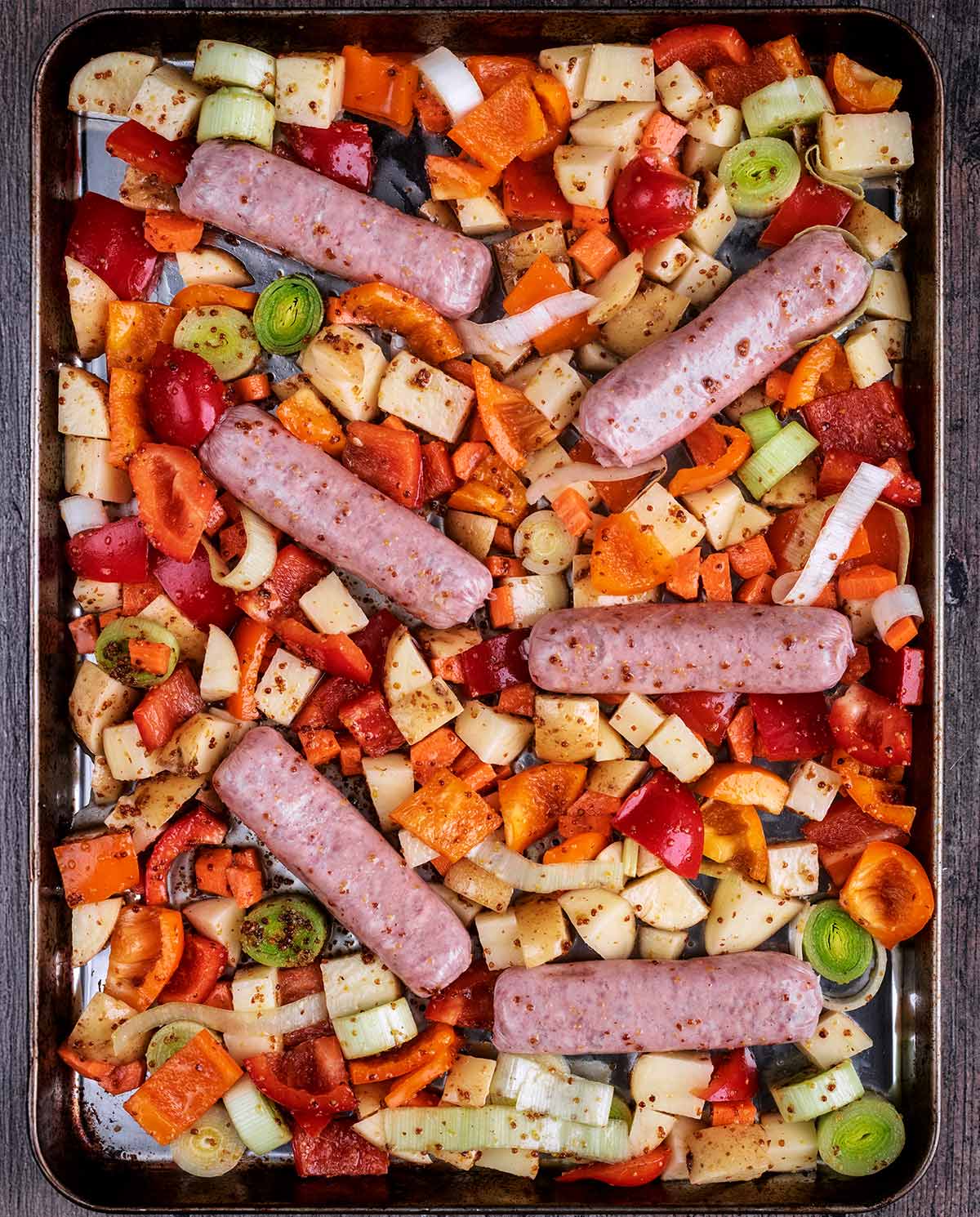 Dressed chopped vegetables and six sausages on a large baking tray.