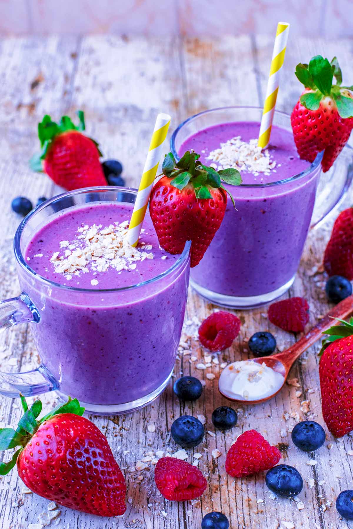 Two purple smoothies in glasses with straws, topped oats and strawberries.
