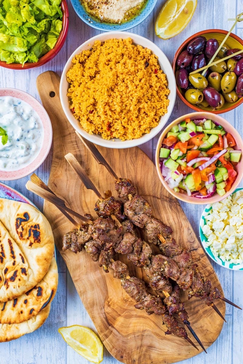 Greek Lamb Souvlaki skewers on a wooden board with couscous, salad, olives and tzatziki.