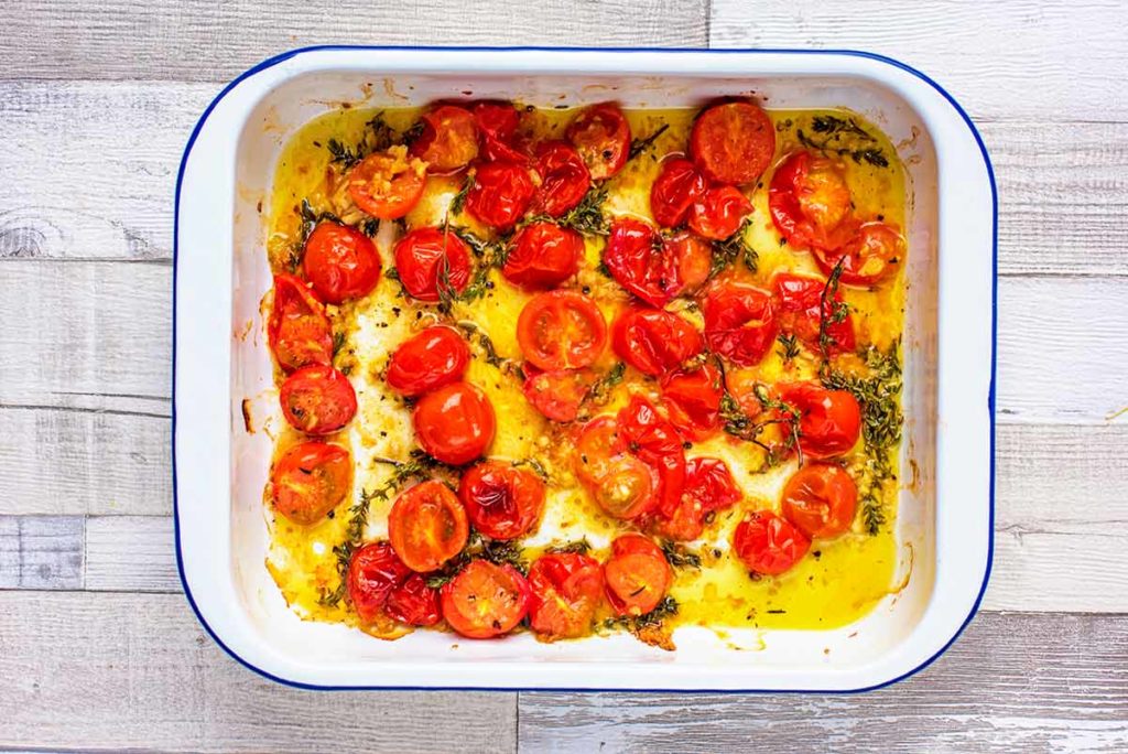 Cooked cherry tomatoes that have burst open in a baking tray with cooked sprigs of thyme.