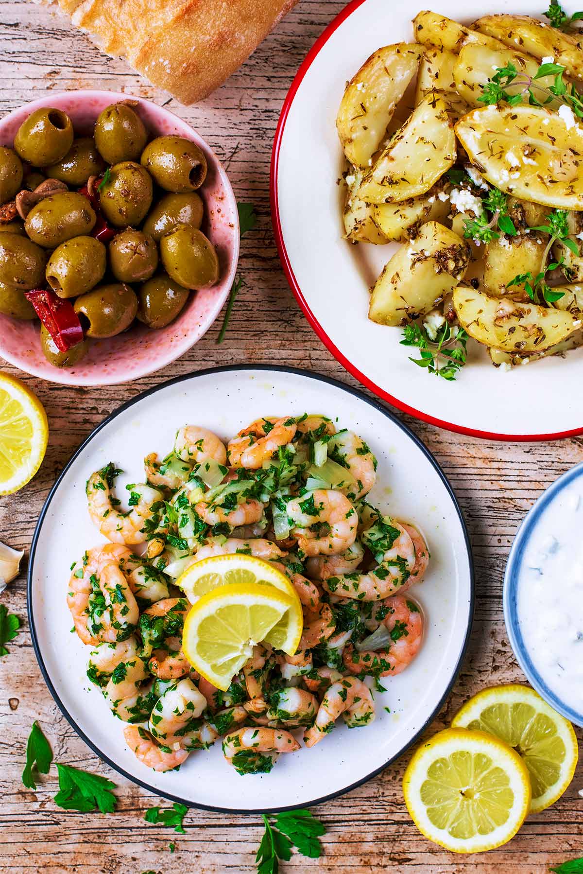 Cooked prawns on a plate next to plates of potatoes, olives and bread.