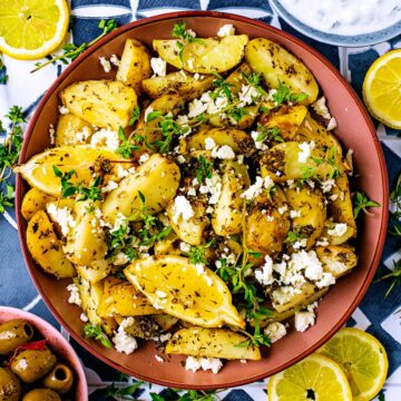 Greek potatoes in a round dish with feta, herbs and lemon wedges.
