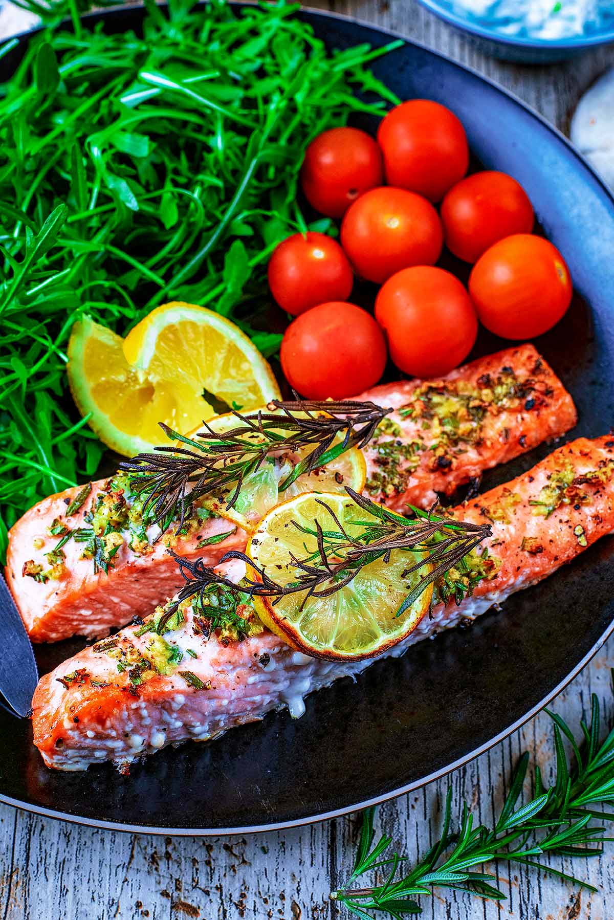 Salmon fillets, lemon wedges, cherry tomatoes and arugula on a black plate.