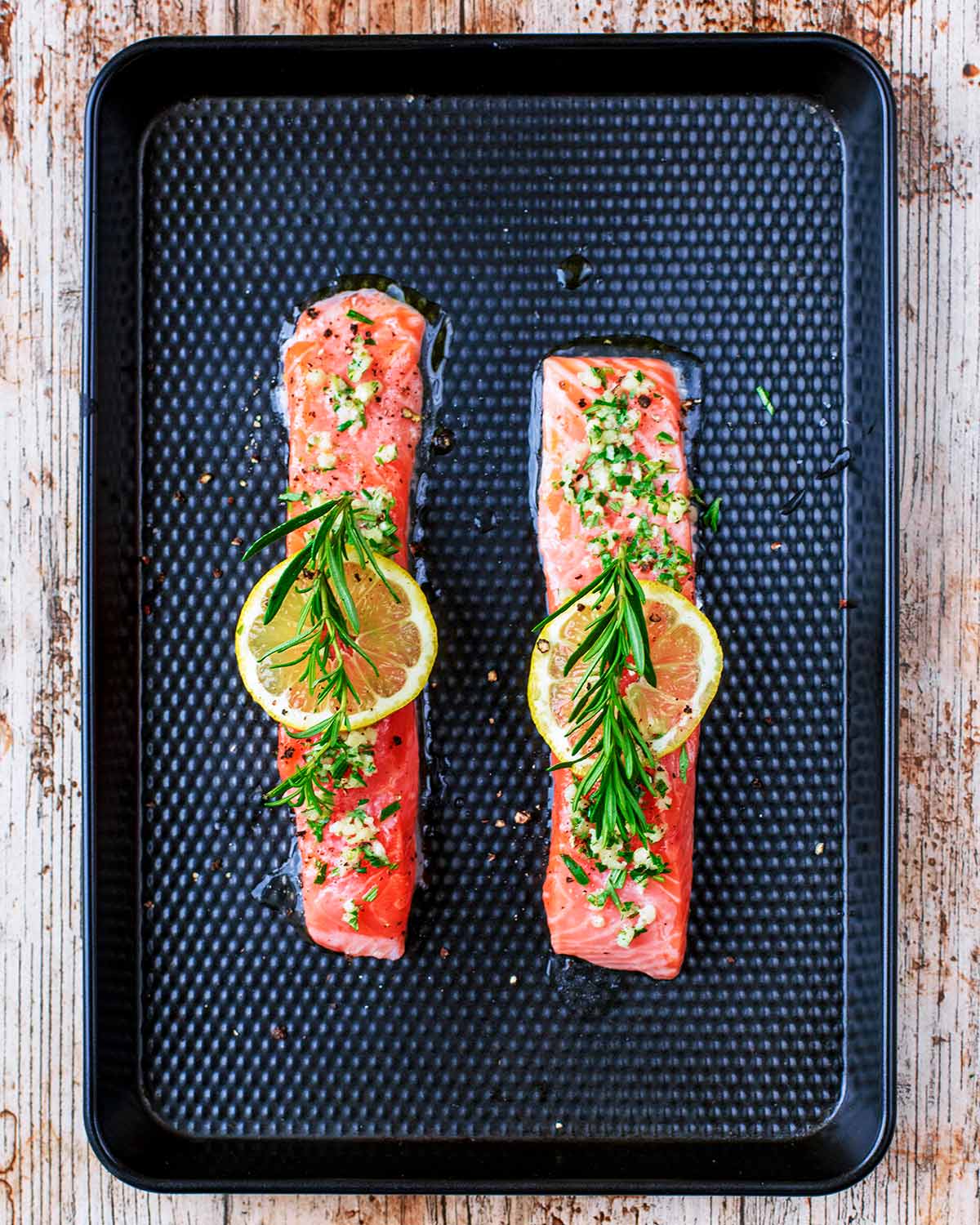 Two salmon fillets on a baking tray with lemon and rosemary