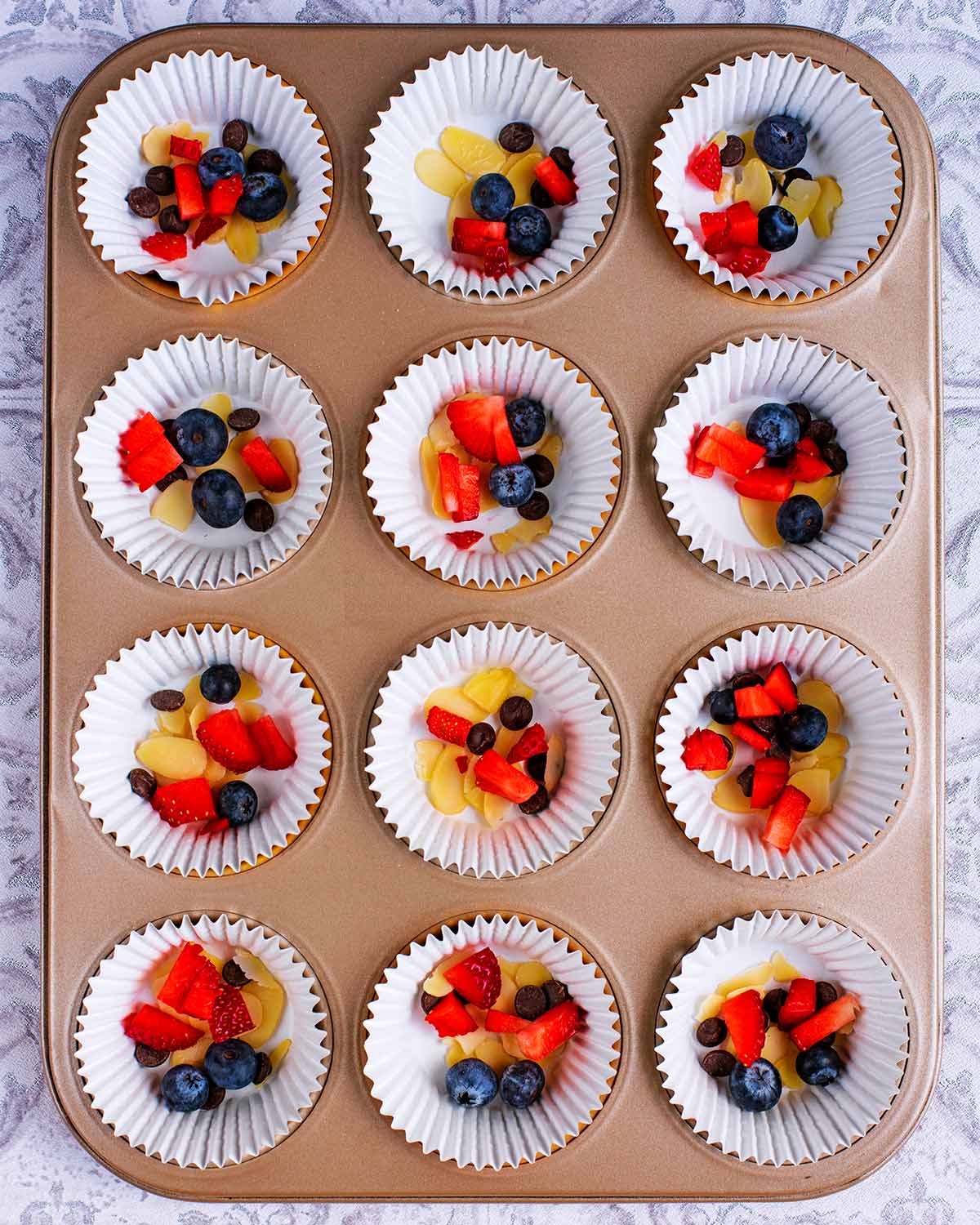 Twelve muffin cases in a muffin tin, each containing blueberries, strawberries and flaked almonds.