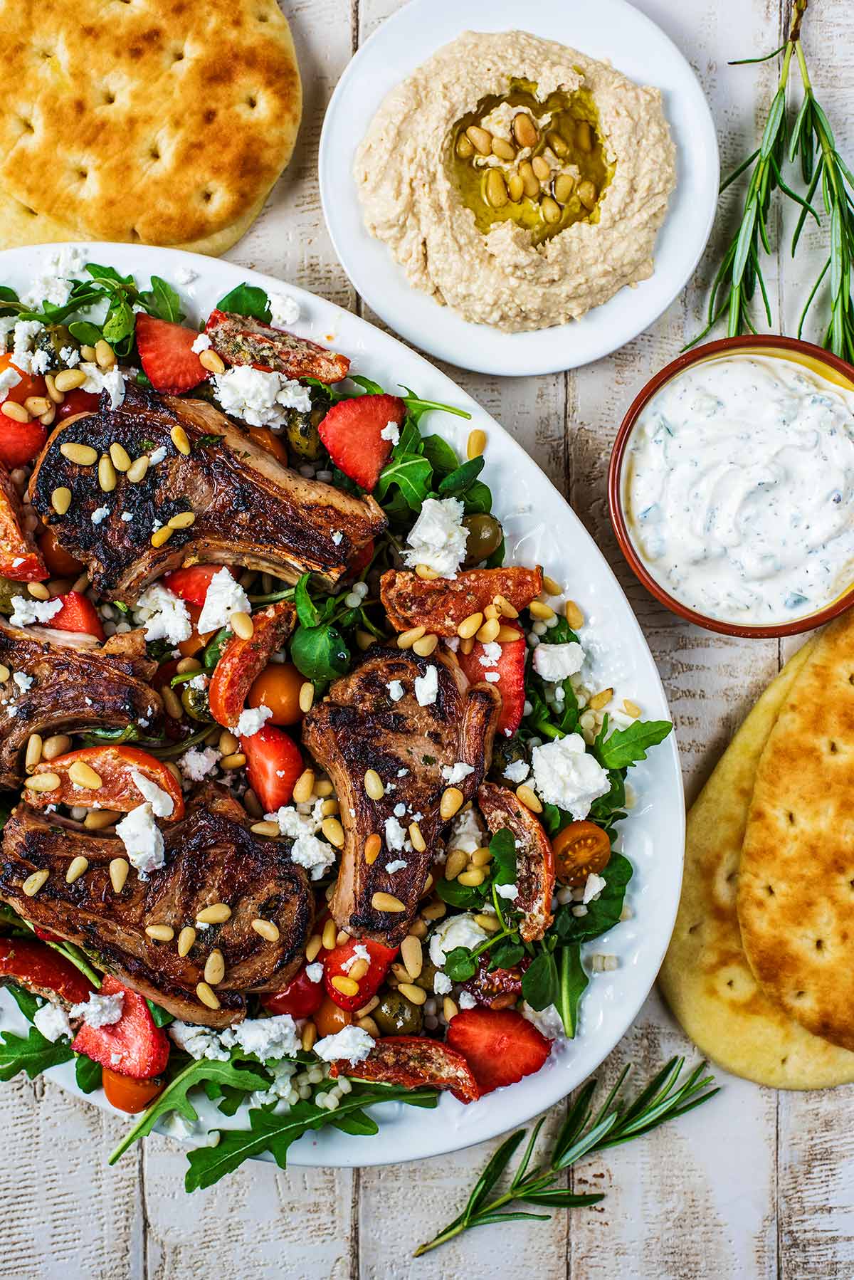 A large white plate of lamb salad next to bread and dips.