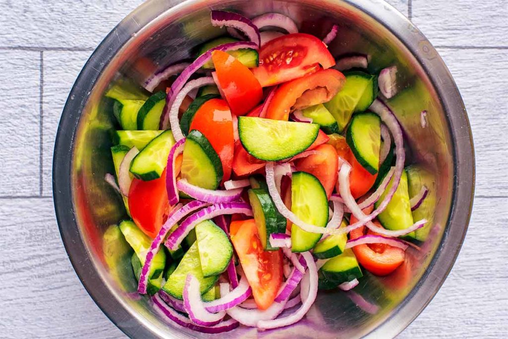 A metal mixing bowl containing sliced cucumber, tomato and red onion.