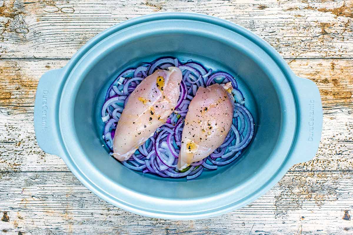 A slow cooker bowl with sliced red onions and two chicken breasts in it.