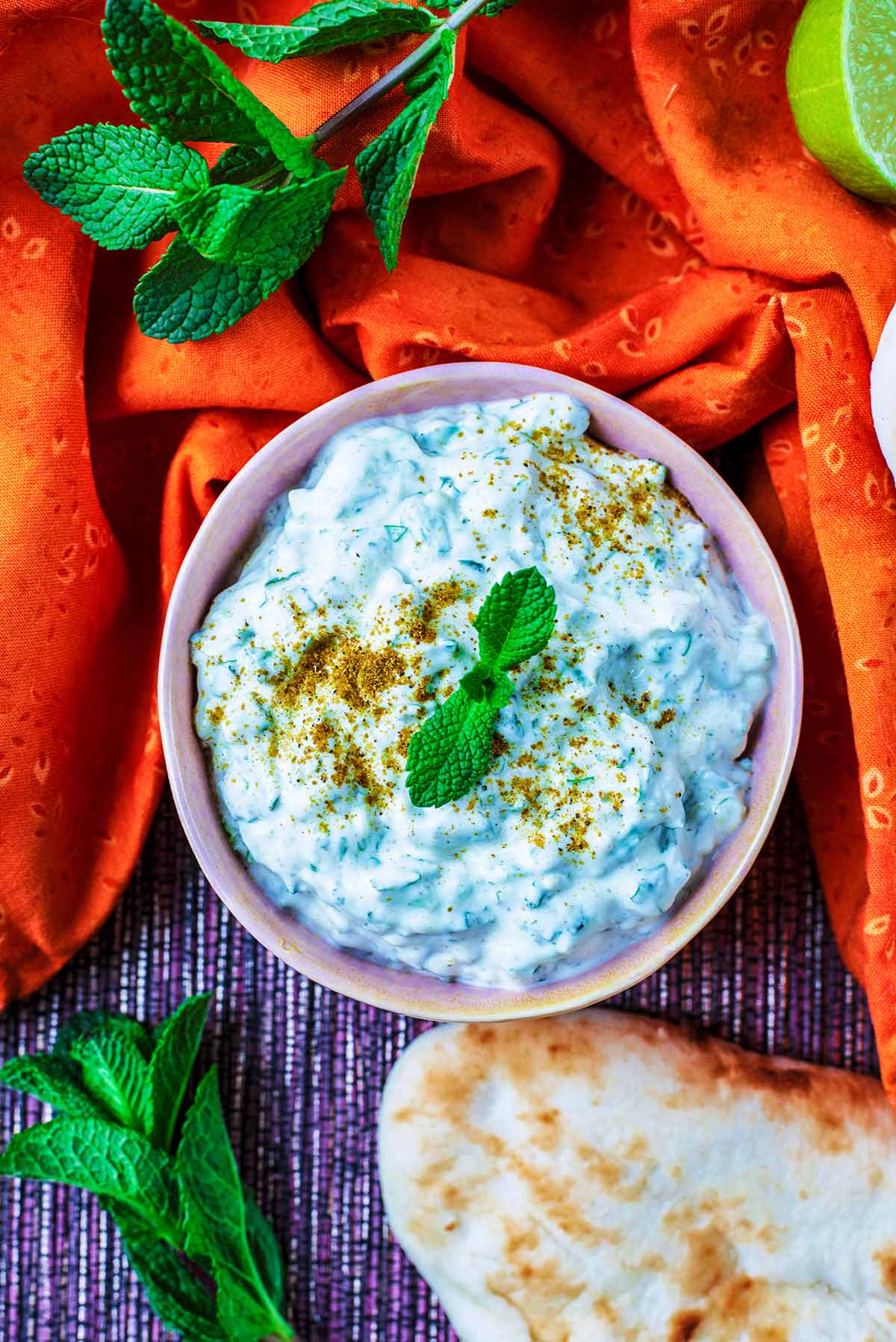 A bowl of raita next to a naan bread and some mint leaves.