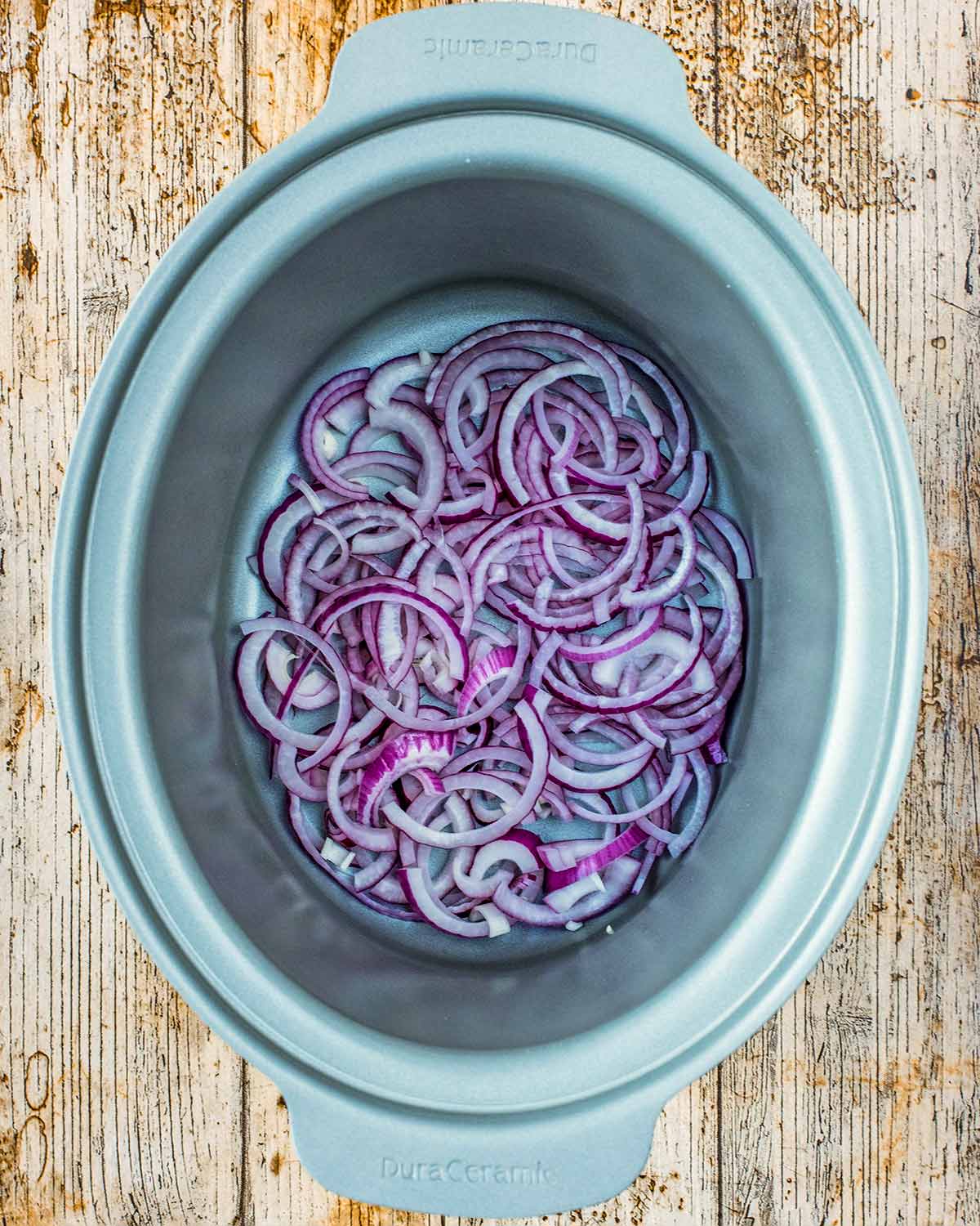 A slow cooker bowl with sliced red onions and two chicken breasts in it.