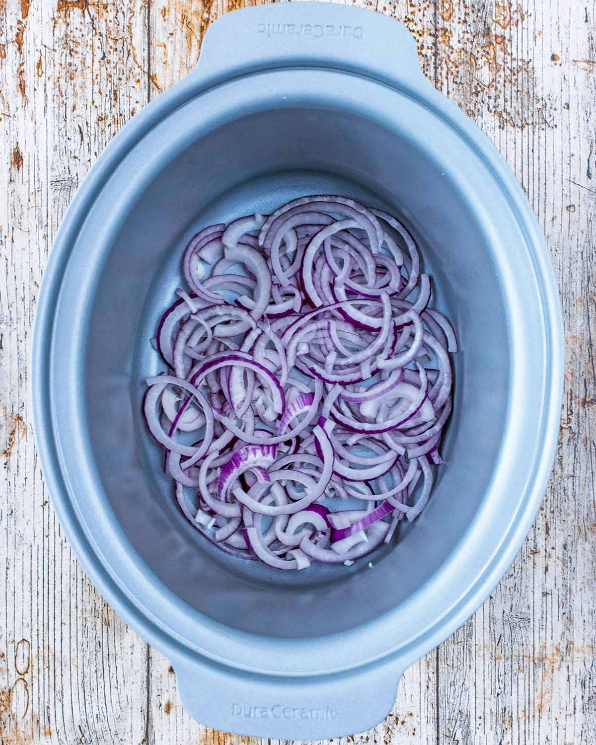 A slow cooker bowl with sliced red onions and two chicken breasts in it.