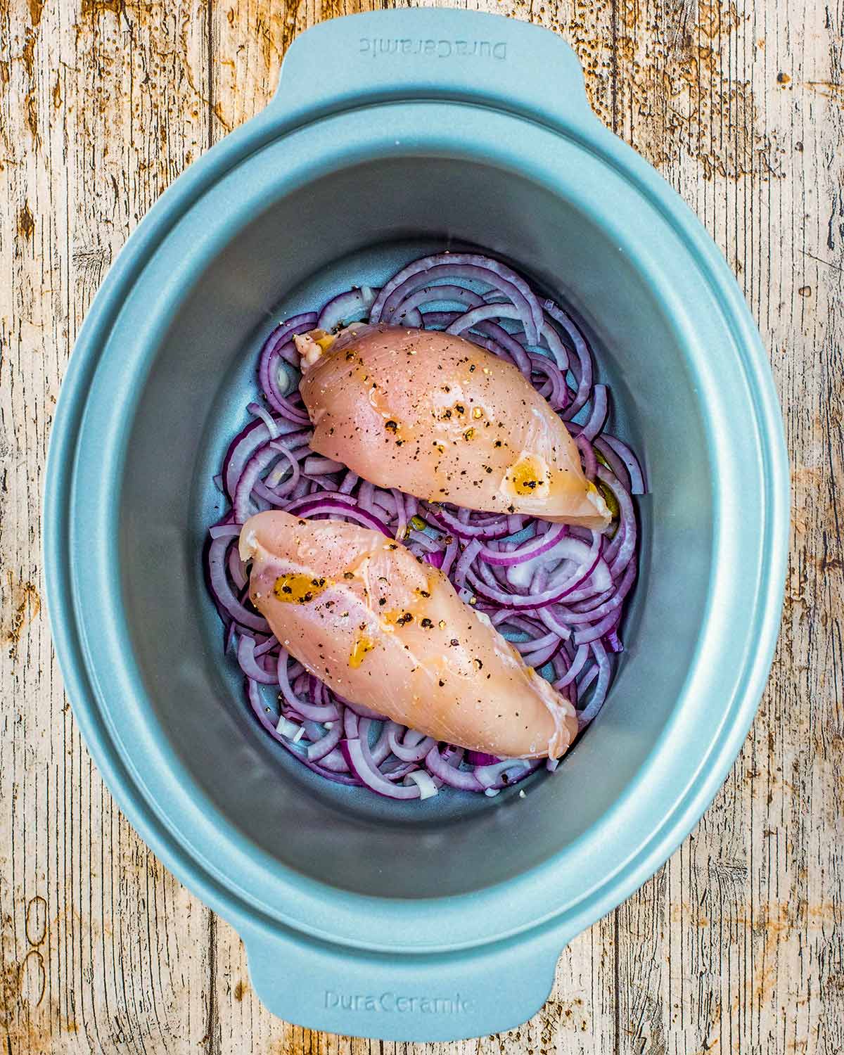 A slow cooker bowl with sliced red onions and two chicken breasts in it.