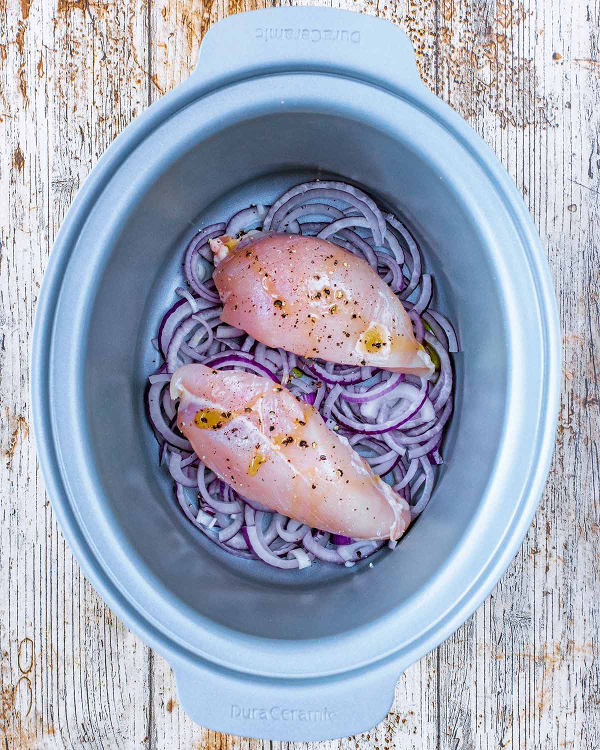A slow cooker bowl with sliced red onions and two chicken breasts in it.
