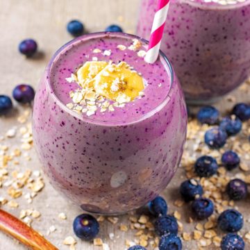 A glass of blueberry smoothie topped with oats and banana. A red and white straw is in the glass and blueberries are scattered around.
