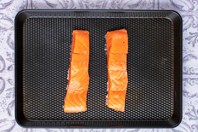 Two uncooked salmon fillets on a black baking tray.