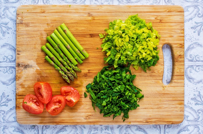 A wooden chopping board with chopped lettuce, asparagus and tomatoes.