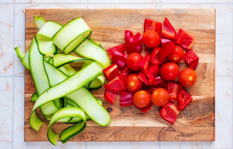 Sliced zucchini, chopped red bell pepper and cherry tomatoes on a wooden chopping board.