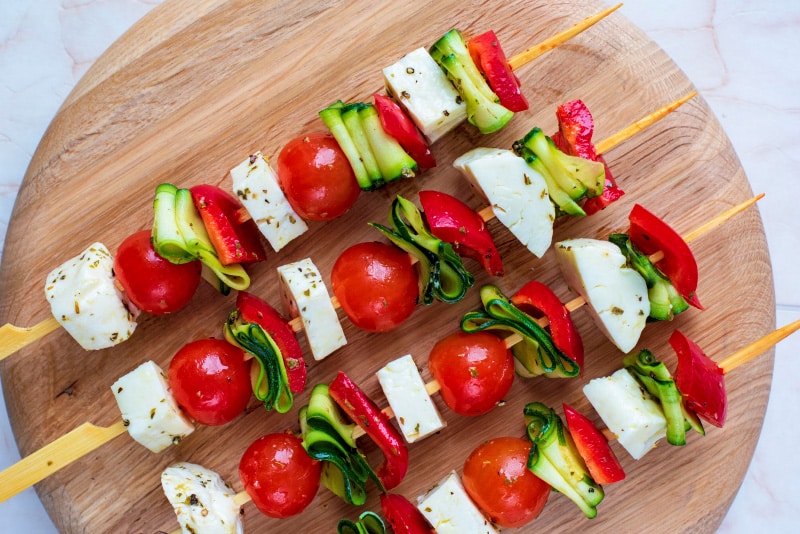 A circular wooden board with vegetable and halloumi skewers on it.
