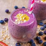 A glass of blueberry smoothie with a red and white straw. Blueberries are scattered around.