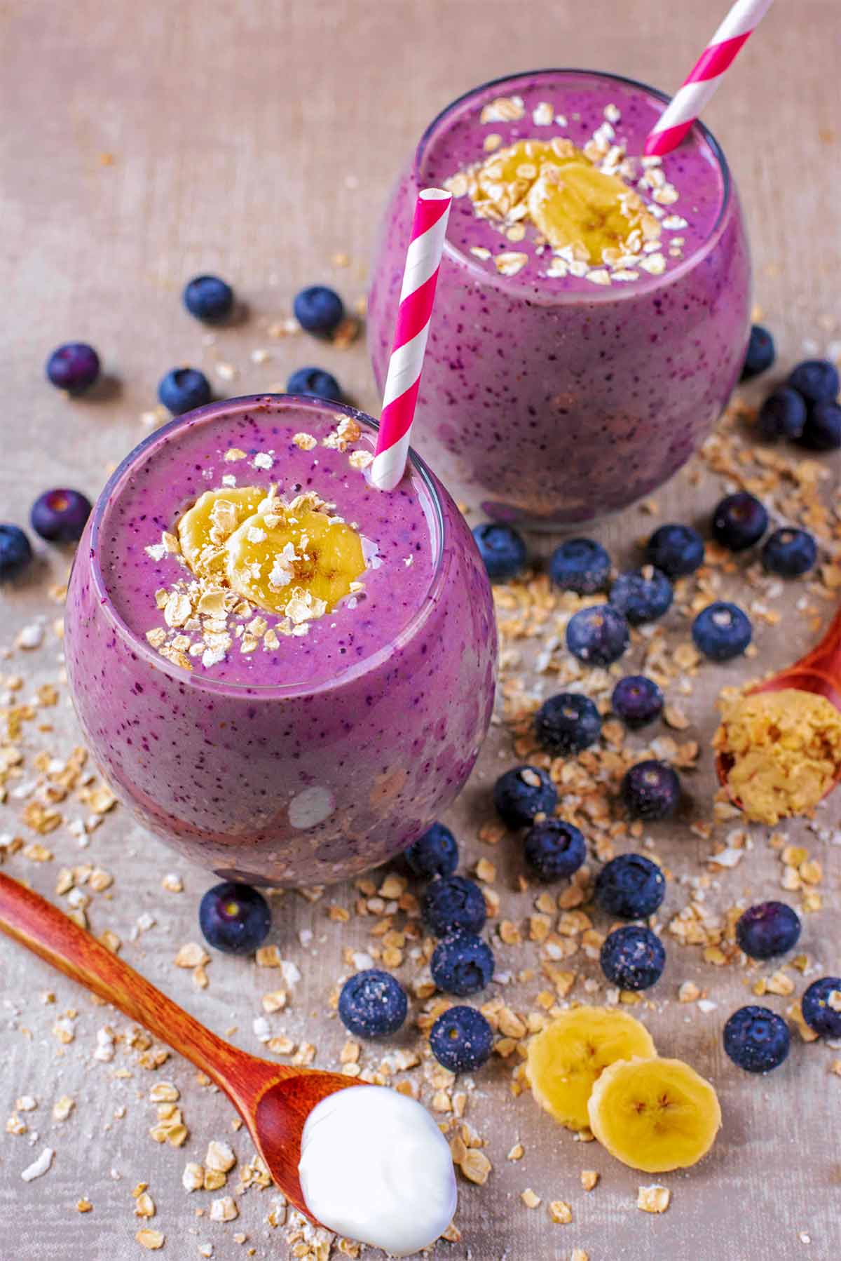 Two glasses of purple smoothie surrounded by blueberries and oats.