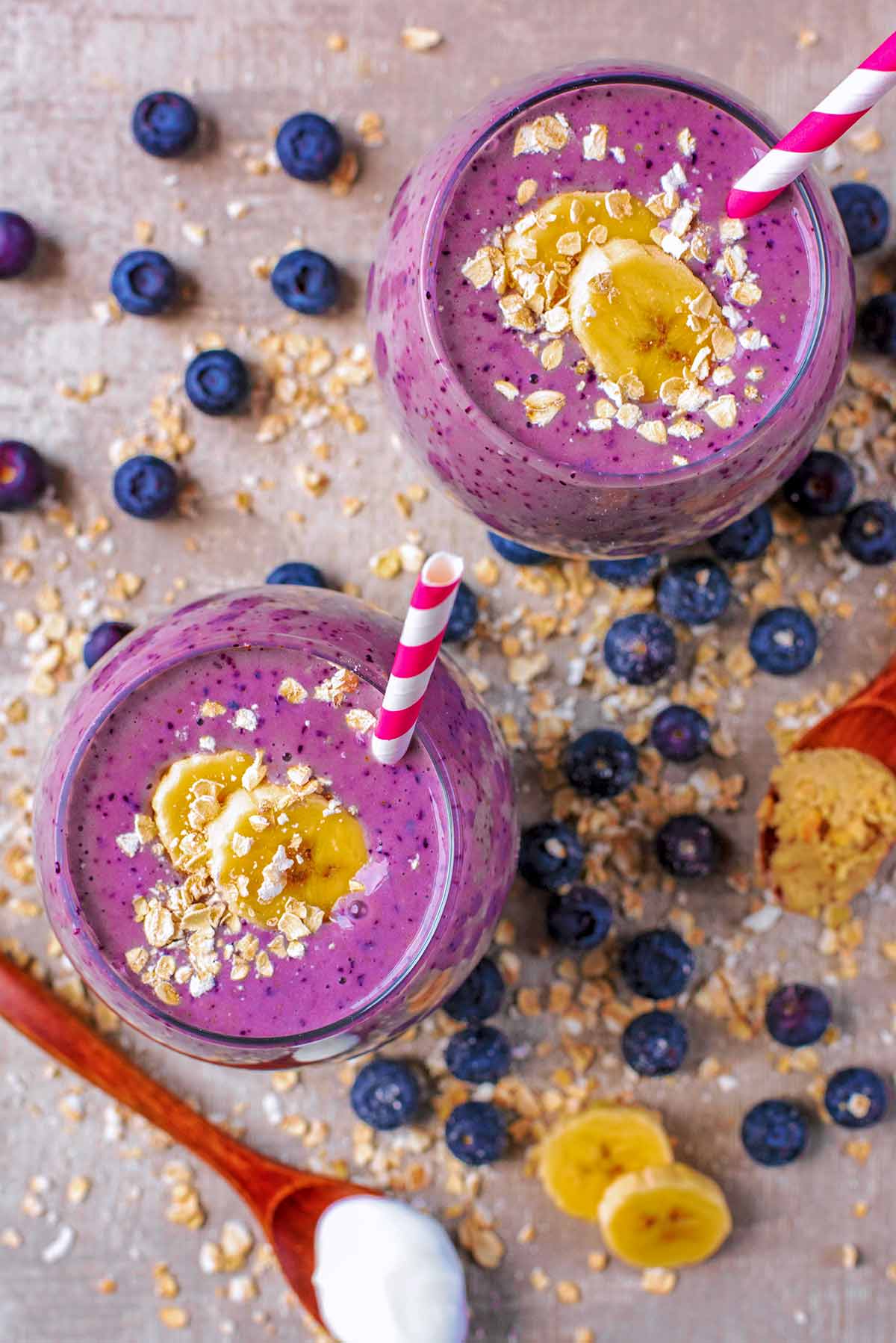 Two smoothies with straws sit amongst blueberries and sliced banana.
