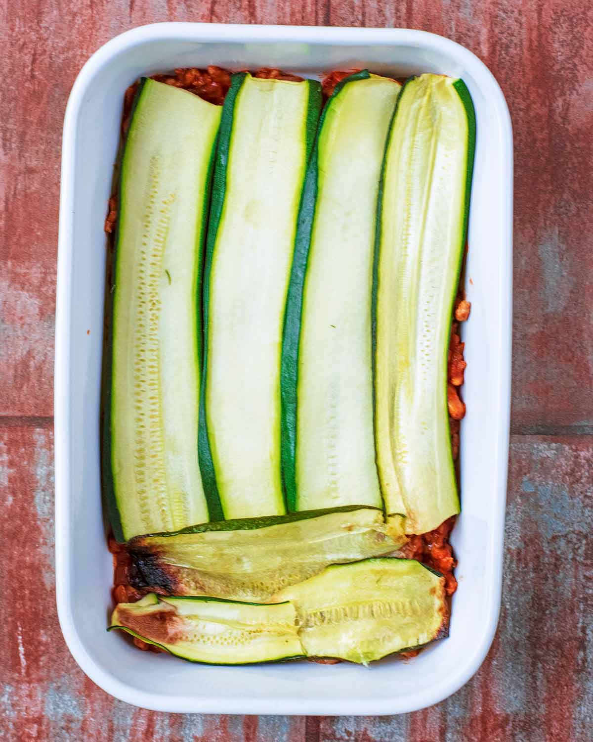 A baking dish containing a tomato meat sauce and strips of cooked courgette strips.