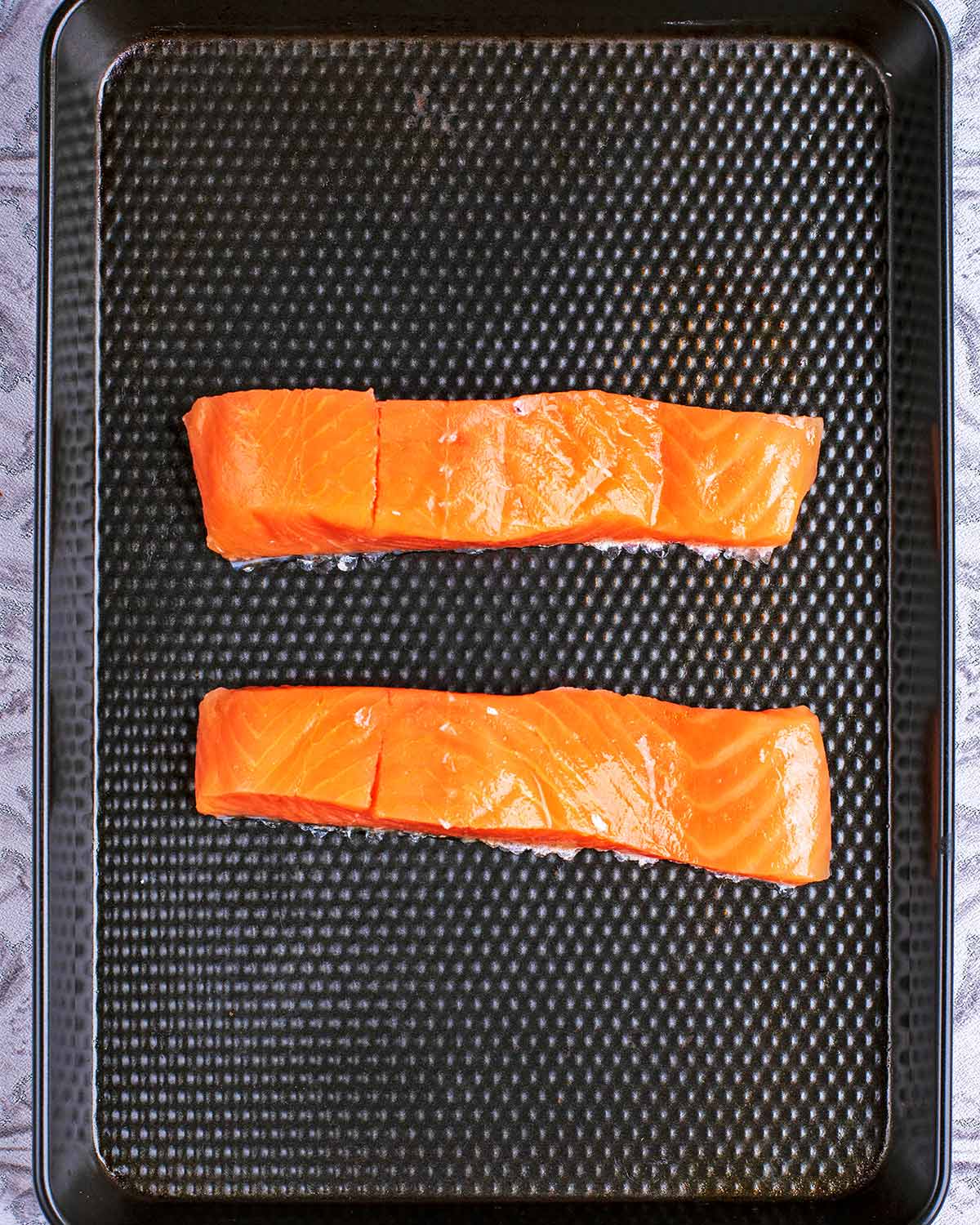 Two uncooked salmon fillets on a black baking tray.