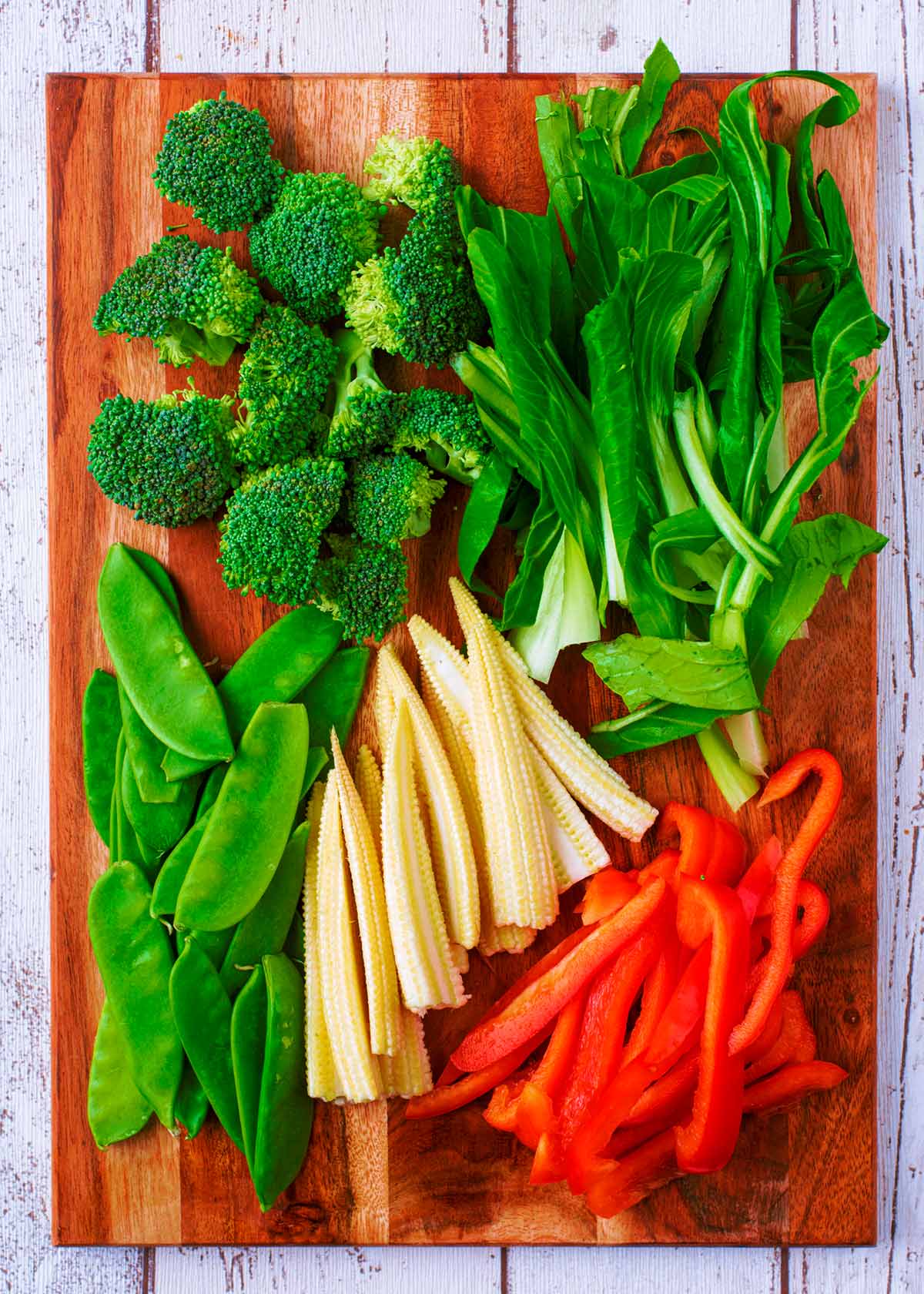 A chopping board with broccoli, pak choi, red pepper, baby corn and mangetout.