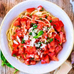 Tomato Basil Pasta in a white bowl next to a tomato, basil leaves and lemon halves.