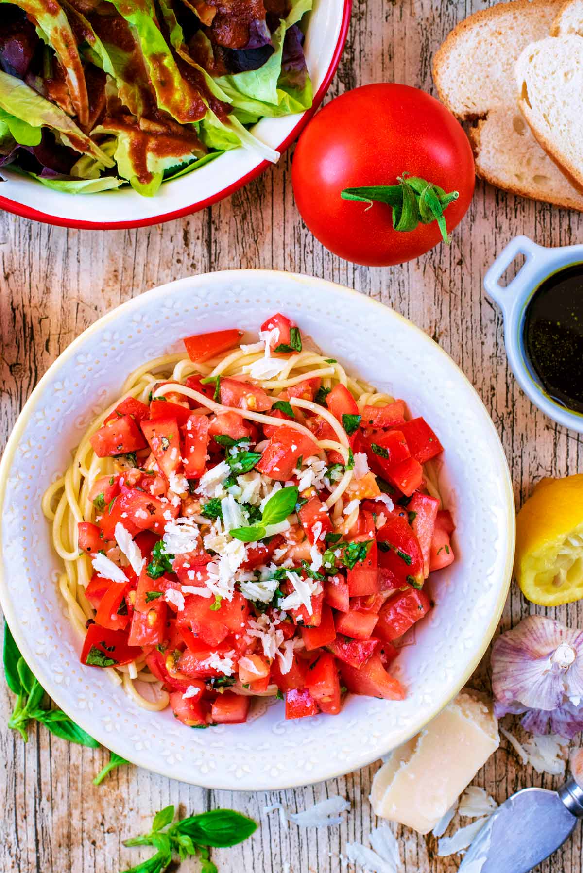 A bowl of Tomato Pasta next to a bowl of salad, a whole tomato and a halved lemon.