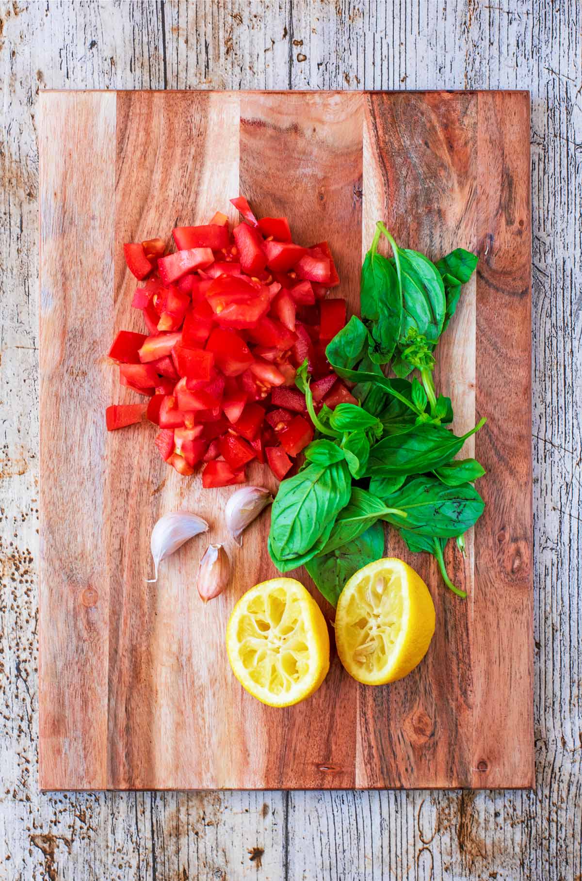Chopped tomatoes, basil leaves, garlic cloves and two lemon halves on a wooden chopping board.