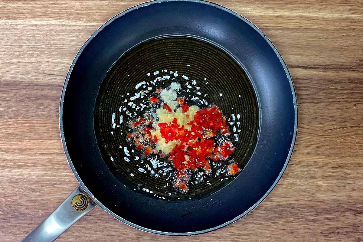 A frying pan with crushed garlic and chopped chilli cooking in it.