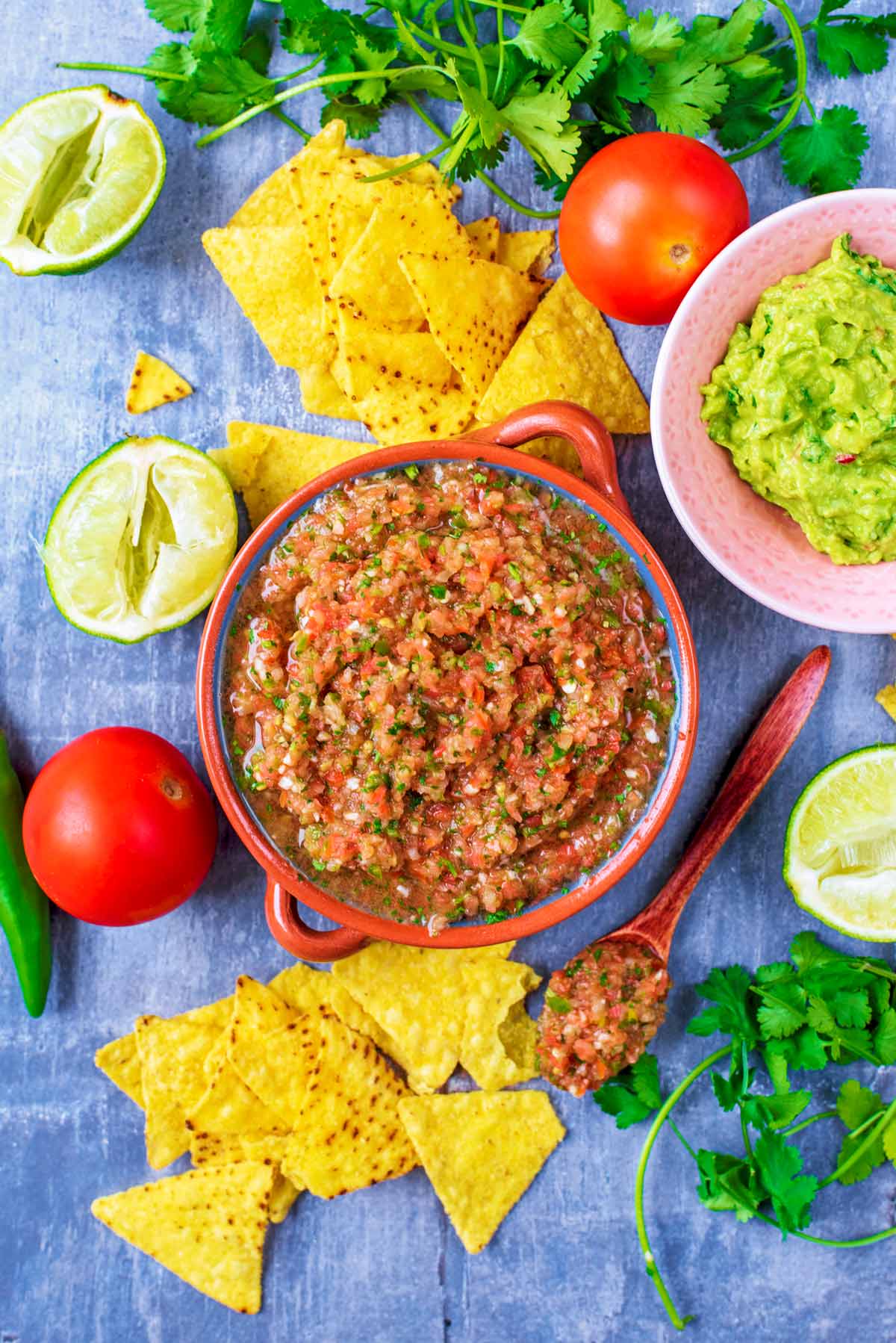 A bowl of Salsa surrounded by tortilla chips, guacamole, limes and herbs.