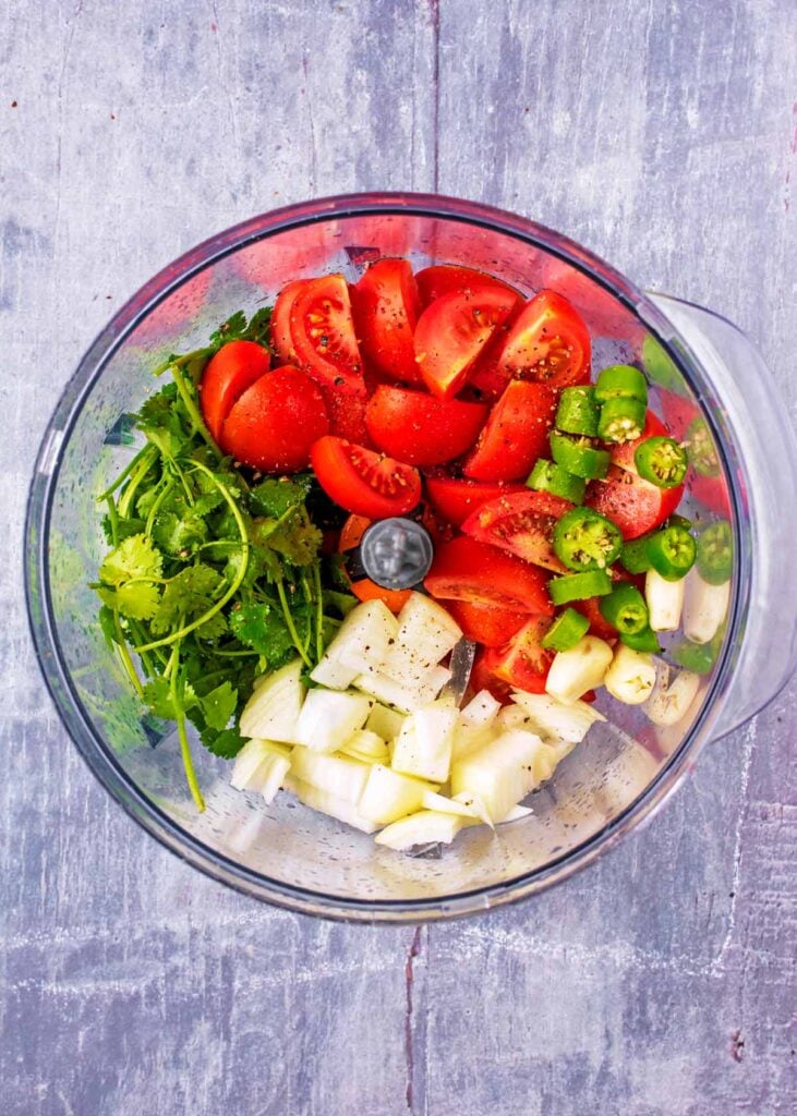 A food processor with chopped tomatoes, onion, chilies, garlic and cilantro.