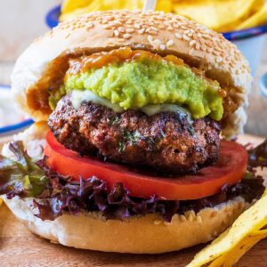 A sesame seed bun containing a Mexican burger, lettuce, tomato, guacamole and relish