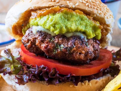 A sesame seed bun containing a Mexican burger, lettuce, tomato, guacamole and relish