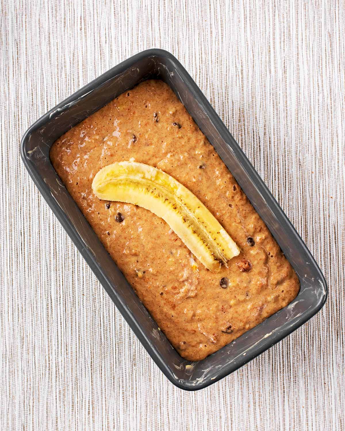 A loaf tin containing a brown cake batter topped with sliced banana.