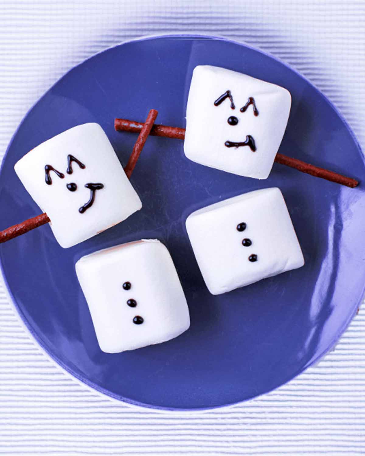 Marshmallows on a plate with icing and chocolate sticks making them look like snowmen.