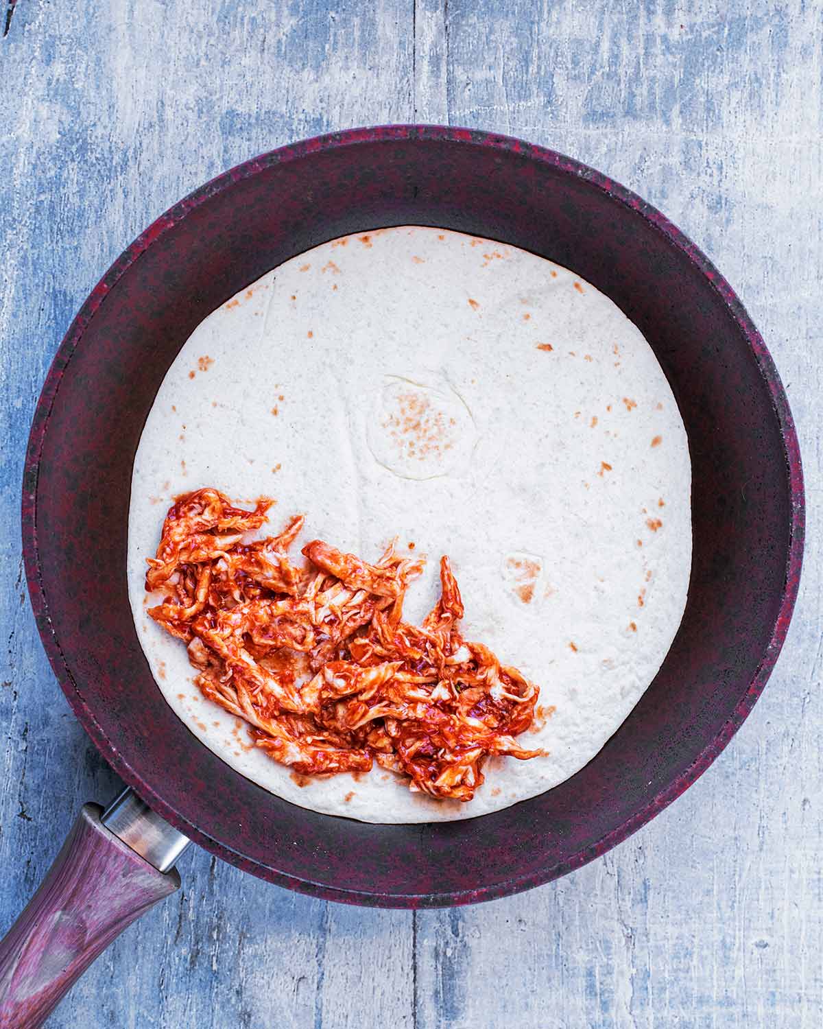A flour tortilla in a frying pan with barbecue chicken on half of it.