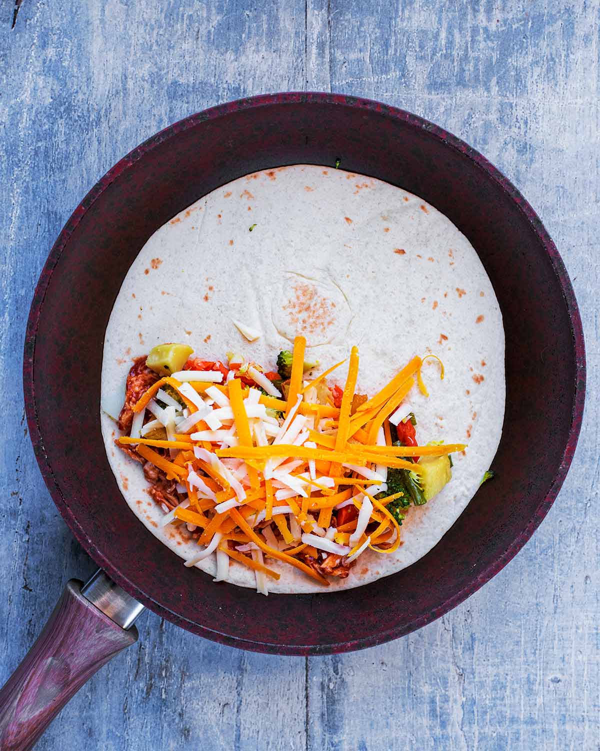 A flour tortilla in a frying pan with barbecue chicken, chopped vegetables and grated cheese on half of it.