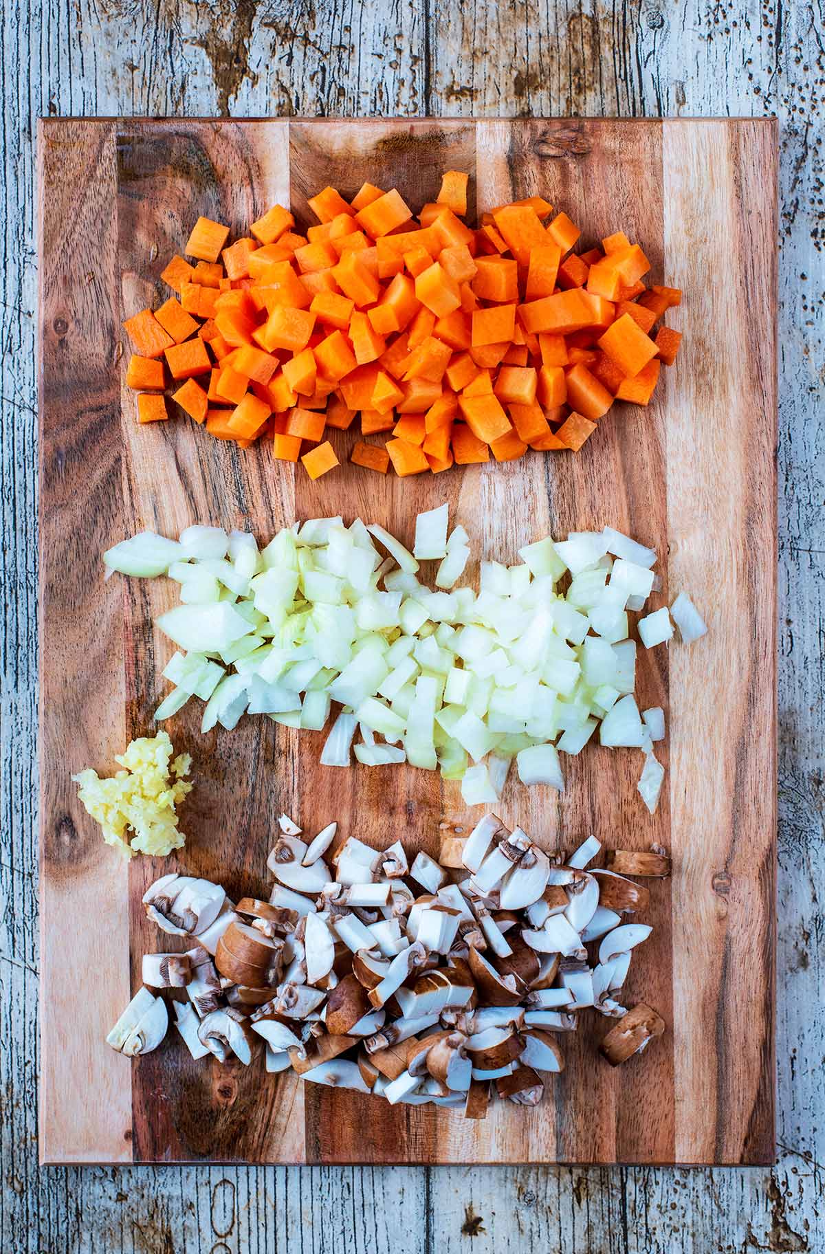 A wooden chopping board with chopped mushrooms, onion, carrot and garlic.