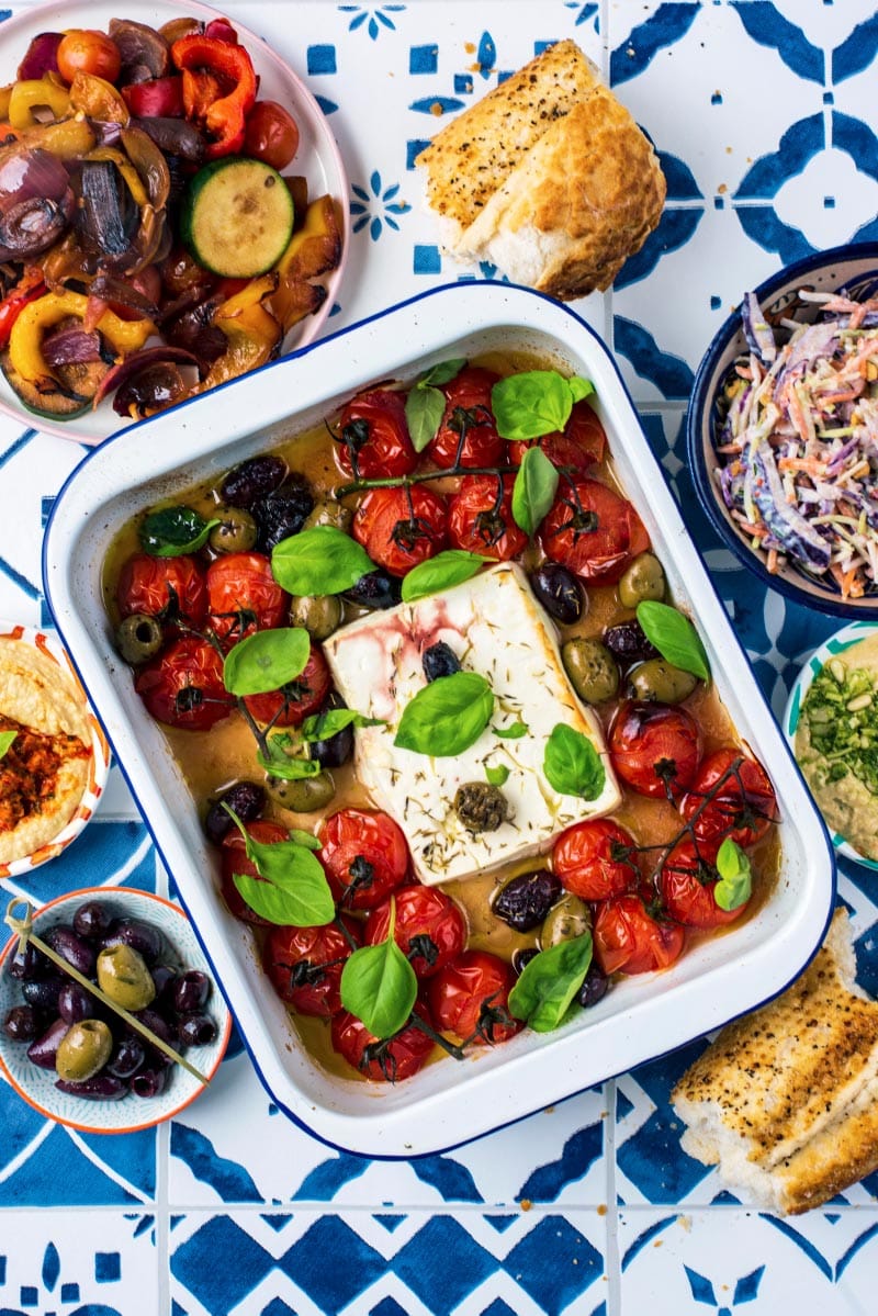 A block of cooked feta in a baking dish with cherry tomatoes, olives and basil leaves.