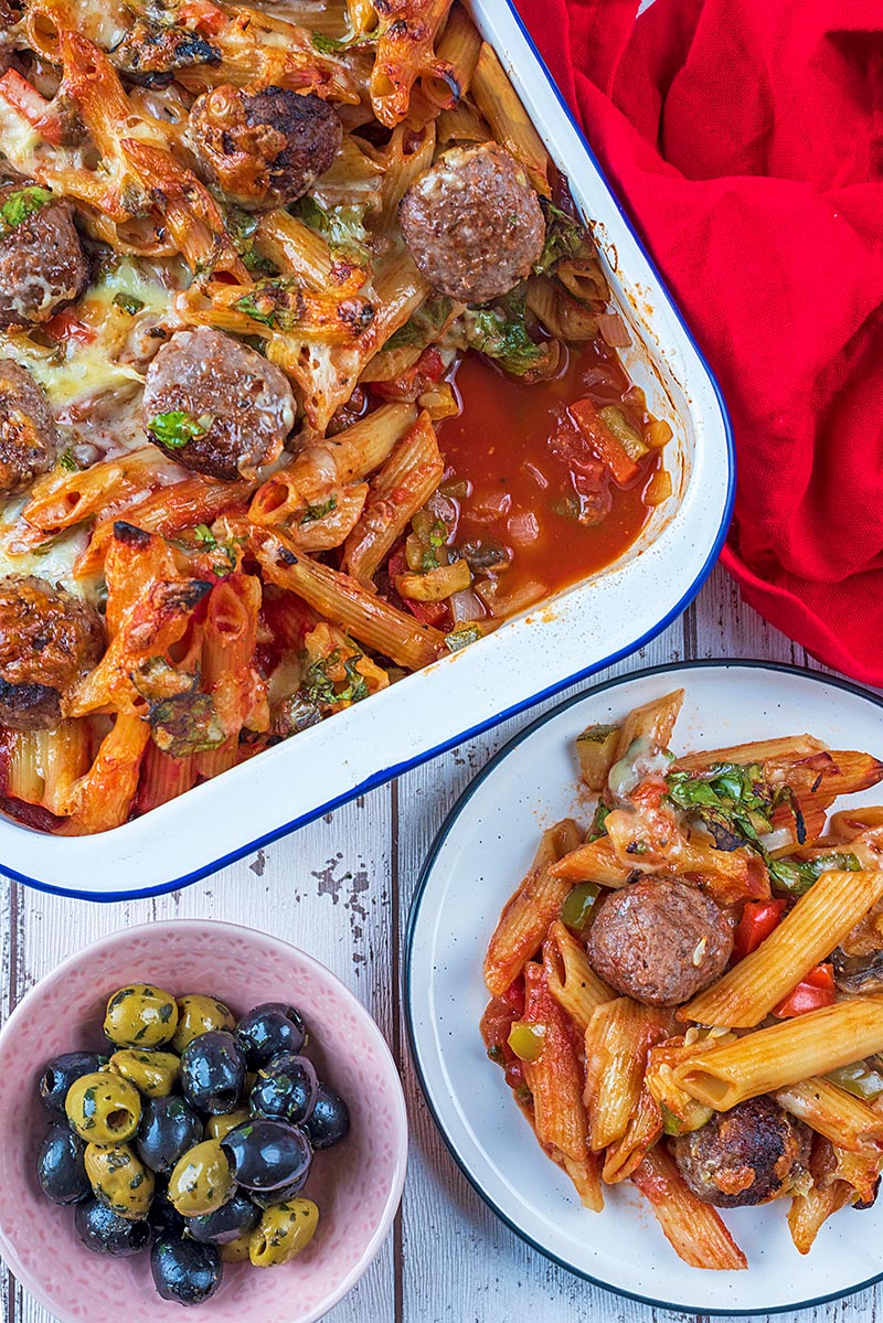 Meatball pasta bake in a large baking dish and on a white plate. A small bowl of olives is next to them